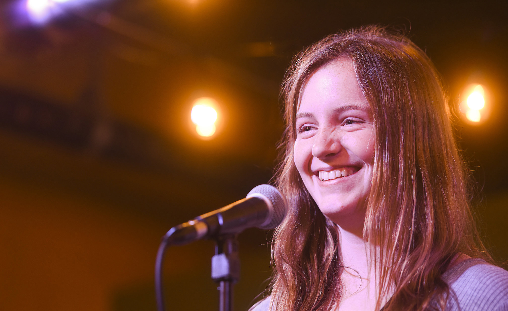 Young adult woman standing at a microphone on stage, she is smiling